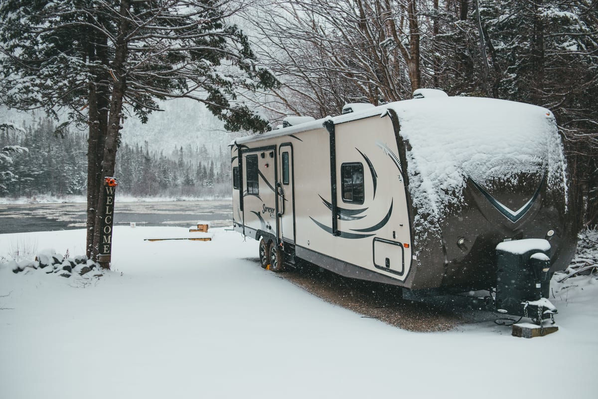 Travel trailer covered with snow placed on shore near river in winter forest in countryside in daylight