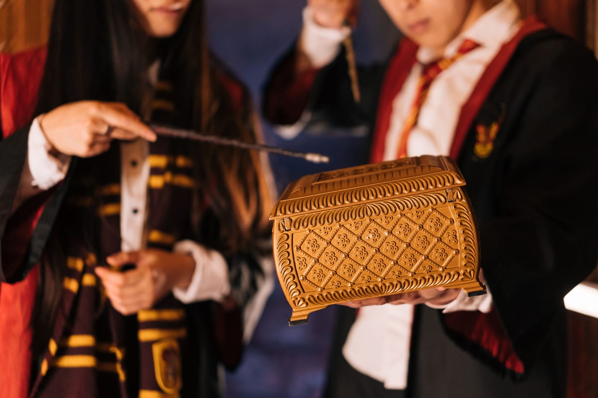 Two people in wizard costumes cast a spell with wands over an ornate box.