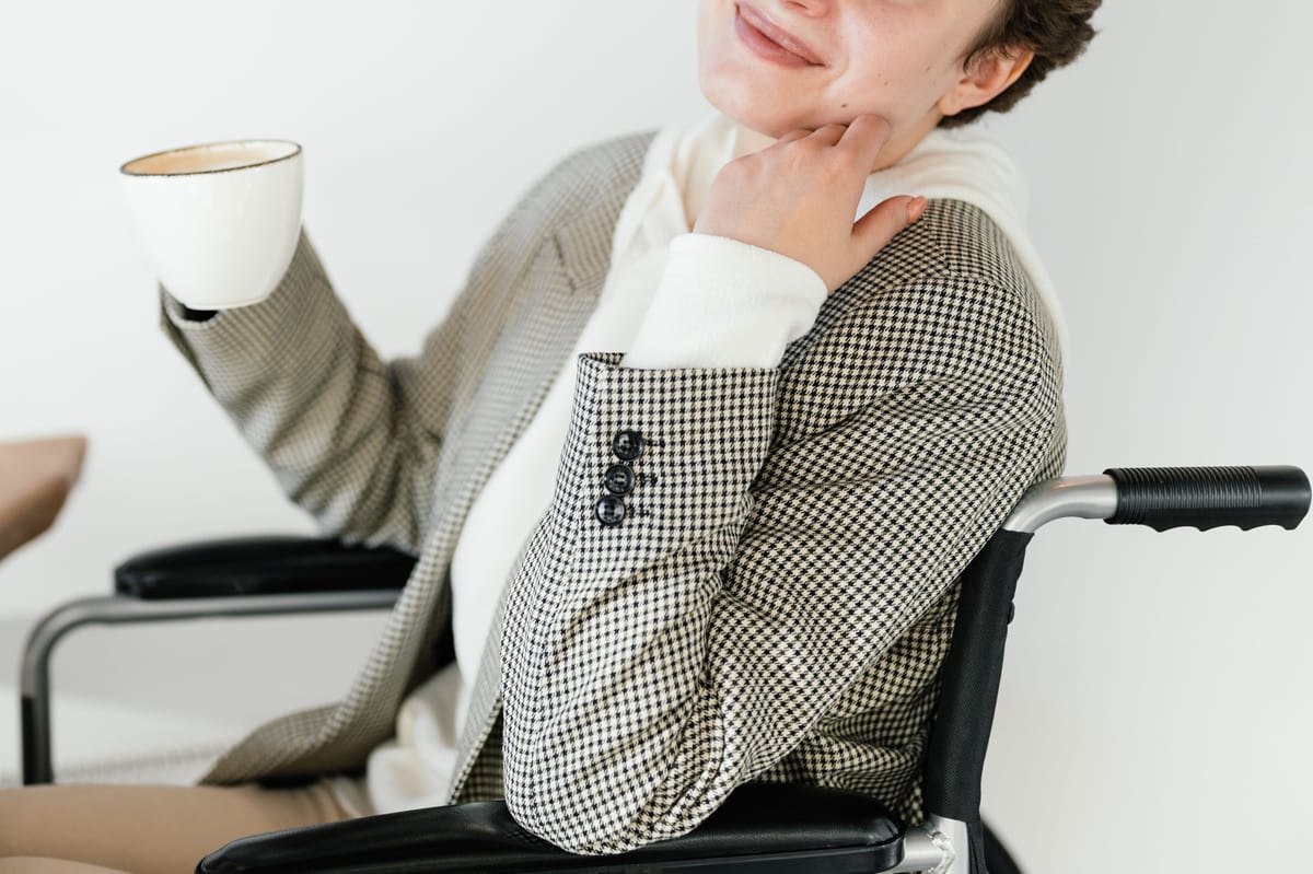 Unrecognizable disabled female in wheelchair with cup of hot drink in hand smiling while spending time in cafe during coffee break