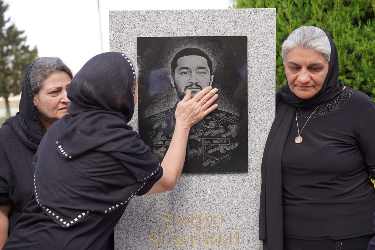 Three women mourning at a memorial stone, touching engraved portrait under daylight.