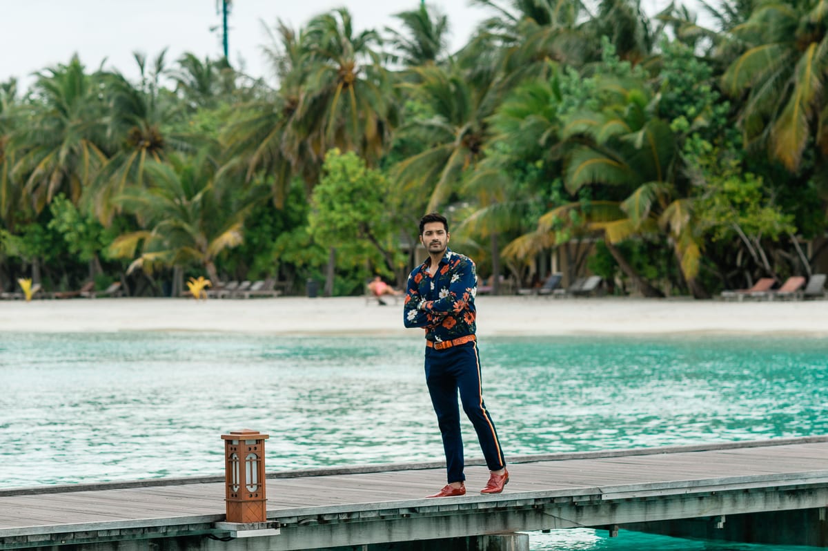 Man posing fashionably on a dock against the picturesque backdrop of Maldives palm trees and turquoise waters.
