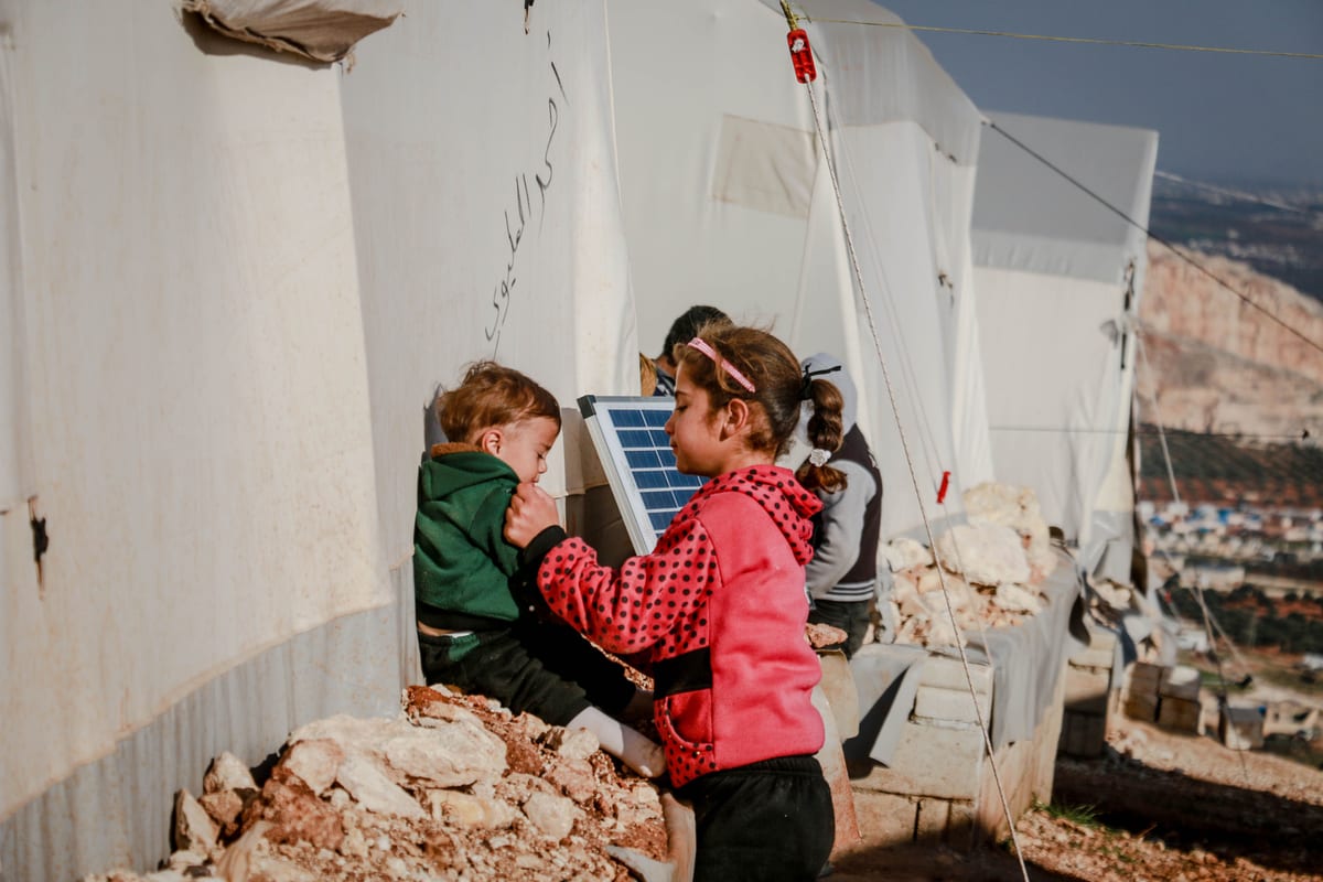 Side view of ethnic girl taking care of brother sitting on stone rubble outside tents in refugee camp