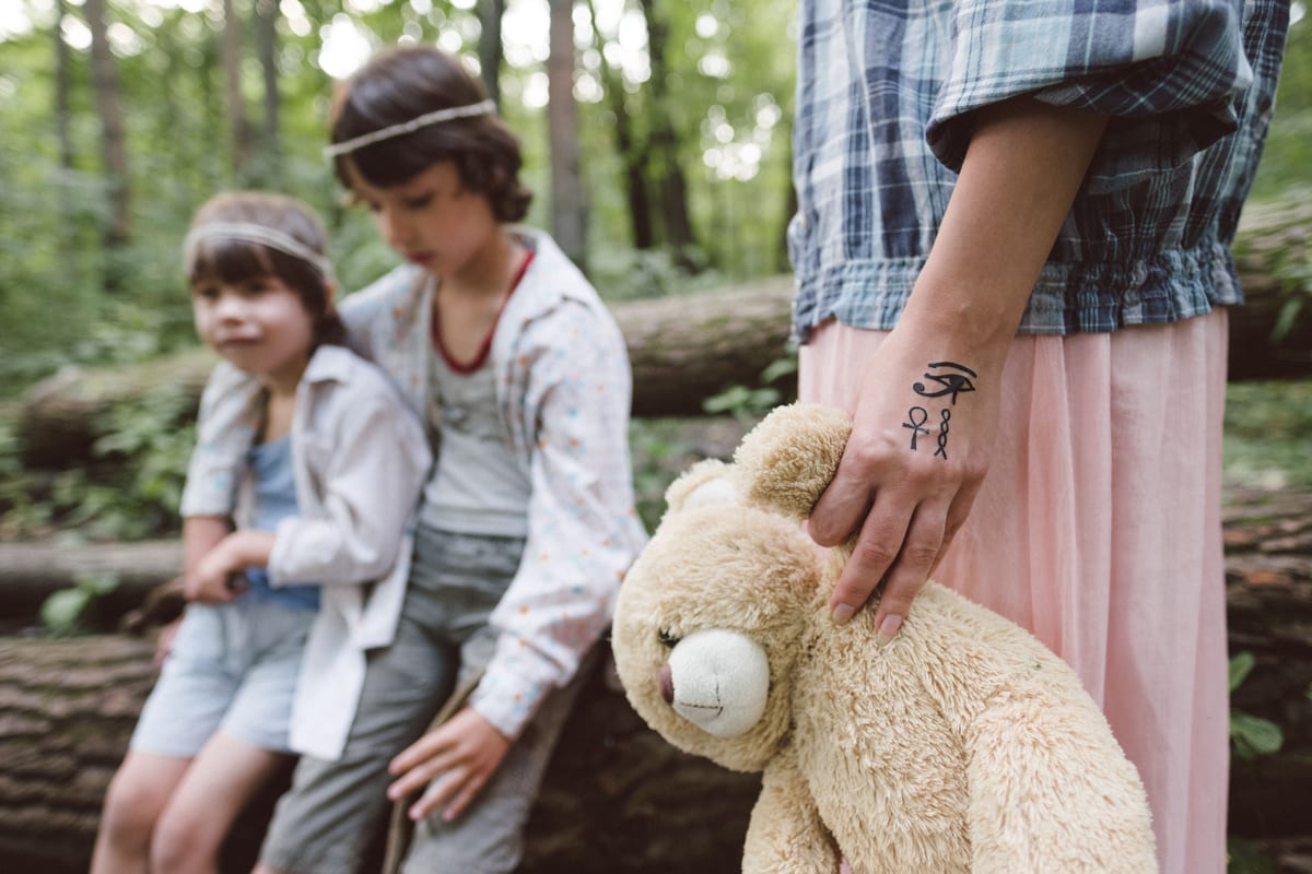 Two children enjoying a playful moment in a forest, accompanied by a teddy bear.