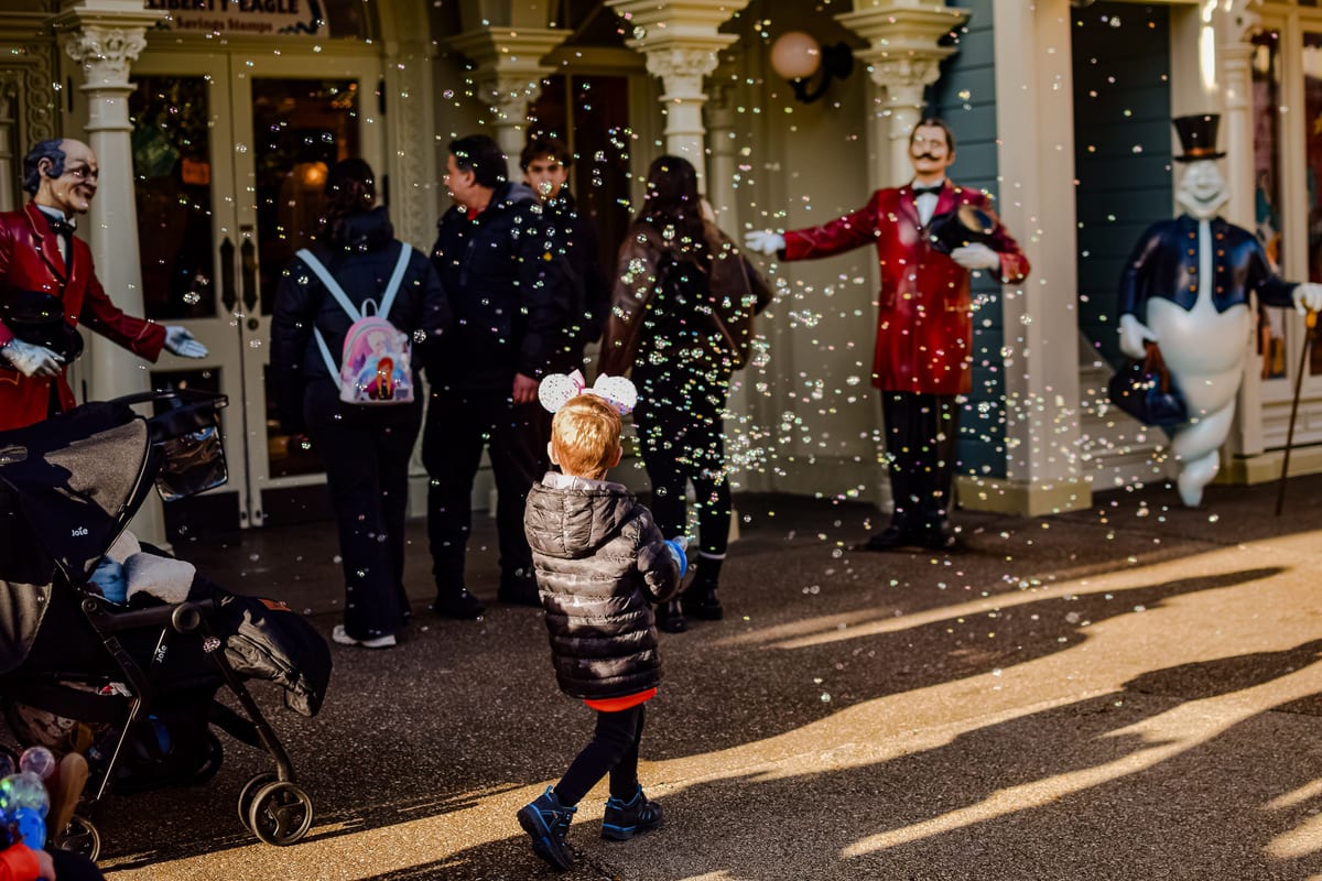 A child wearing mouse ears enjoys bubbles at an amusement park entrance.