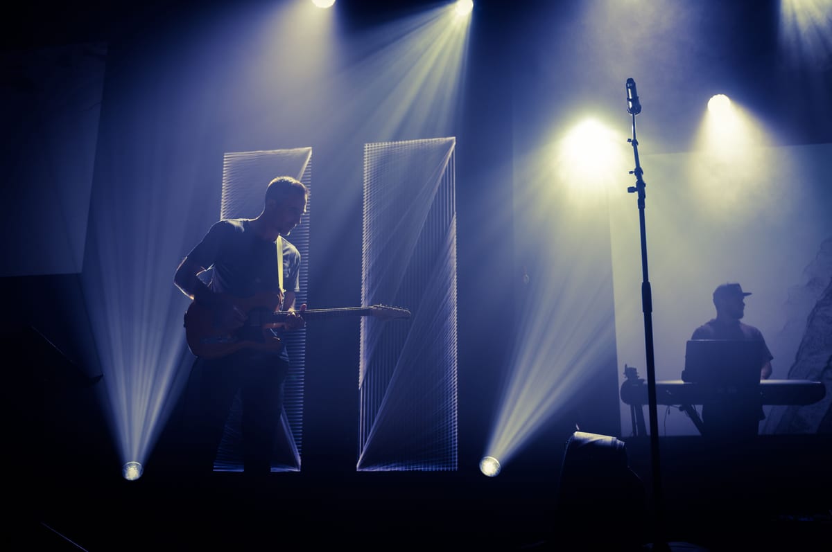 Anonymous male musician performing melody with guitar on stage near microphone and DJ in dark club with spotlights