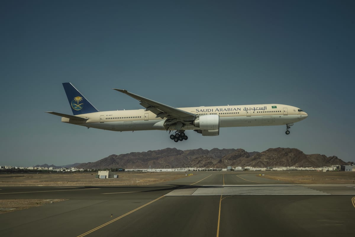 A Saudi Arabian Airlines Boeing 777 landing on a runway with mountainous backdrop.