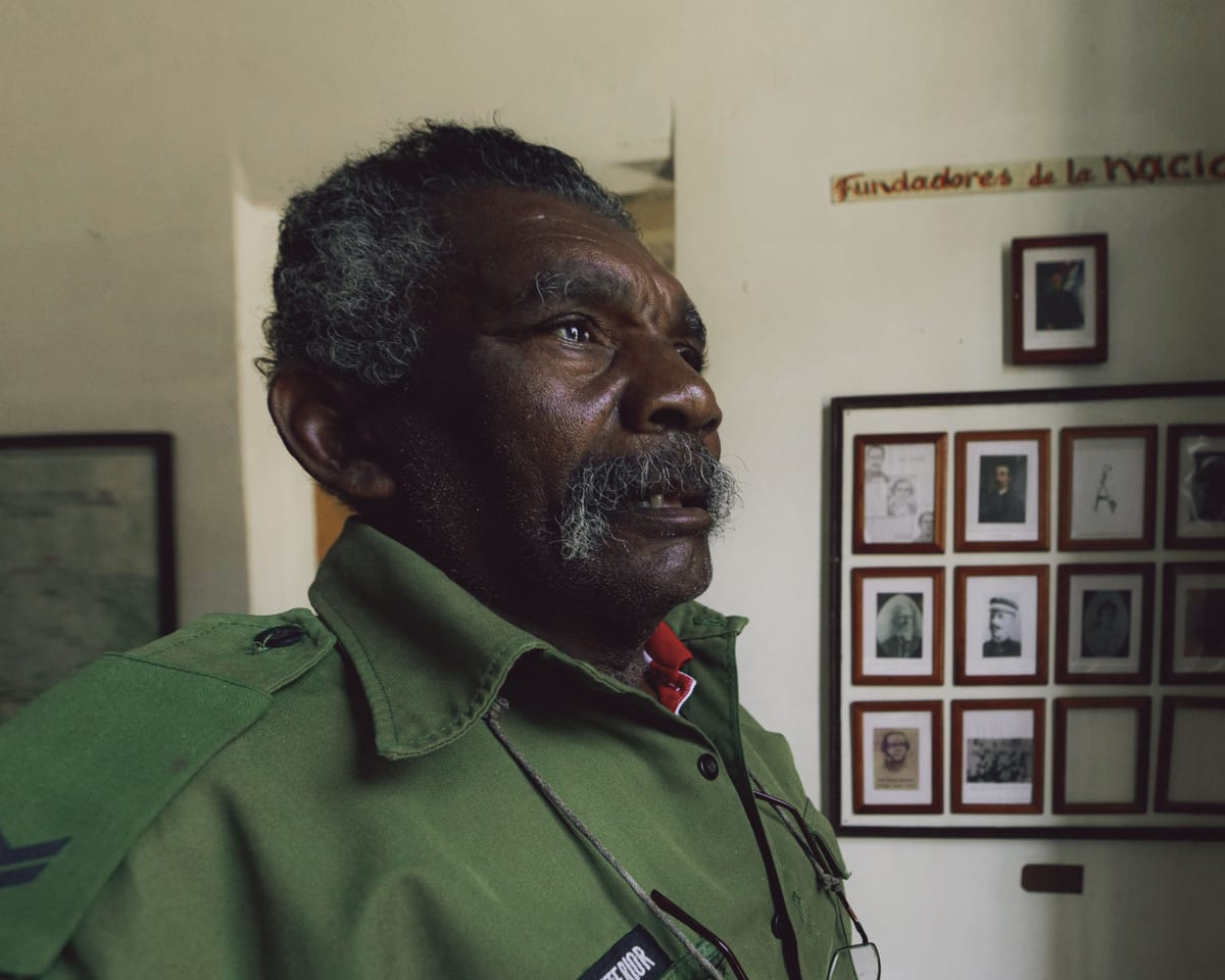 An elderly man with a mustache stands indoors in a uniform, surrounded by framed photos.