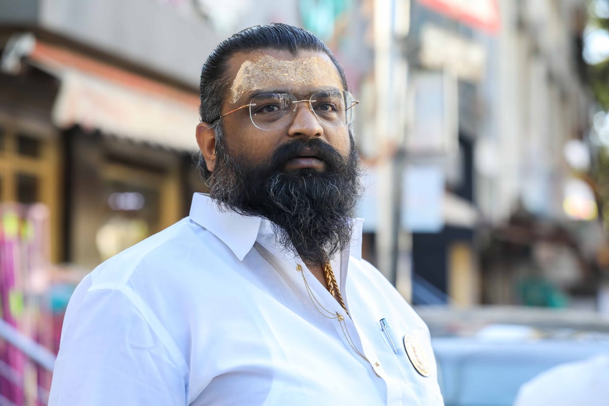 Close-up portrait of a bearded man wearing glasses and a white shirt, captured in an urban outdoor setting.