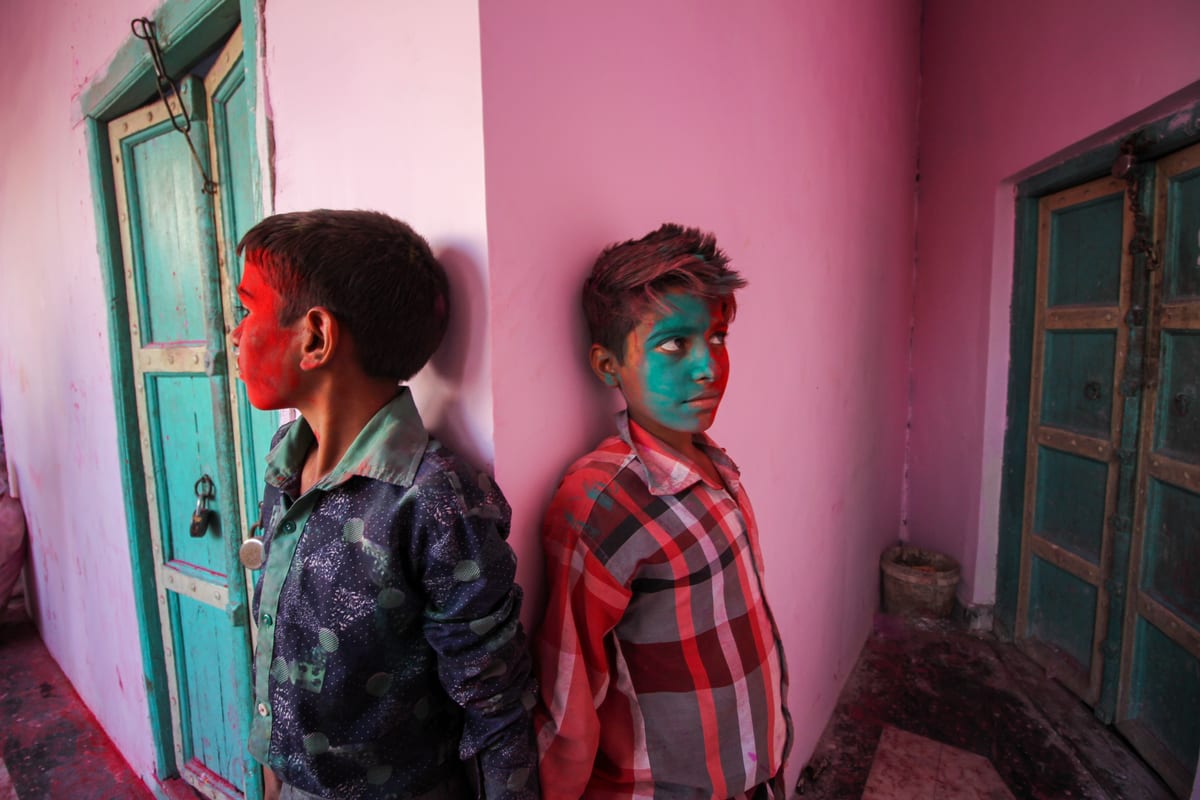 Colorful celebration of Holi Festival with two boys covered in vibrant face paint leaning against a pink wall.