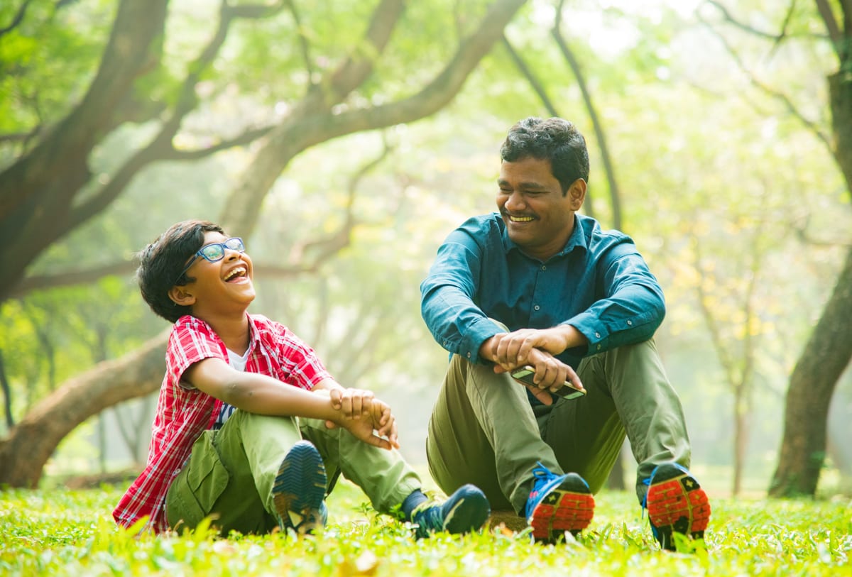 A joyful moment between father and son sitting in a Bengaluru park, sharing laughter.