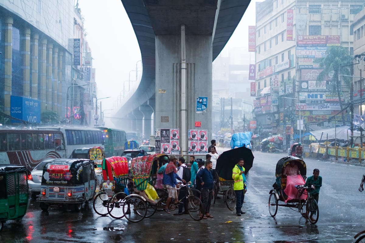 People and rickshaws navigate a busy city street under an overpass during a rainy day.