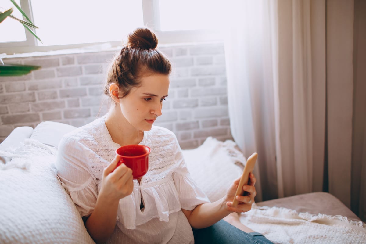 Woman relaxing at home with coffee in hand, browsing her phone comfortably.