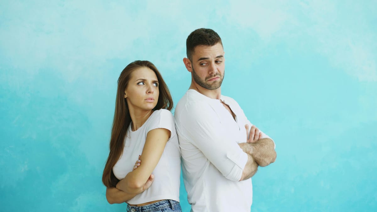 Young couple stands back to back with crossed arms against blue background.