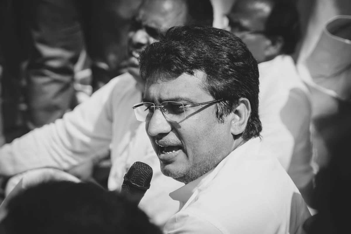 Black and white image of a man with eyeglasses speaking into a microphone at an event.