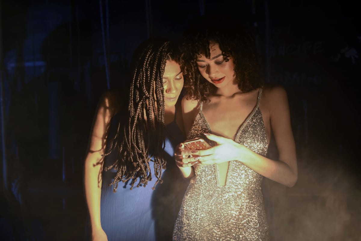 Two fashionable women in evening dresses sharing a moment on a night out, illuminated by a glowing smartphone screen.