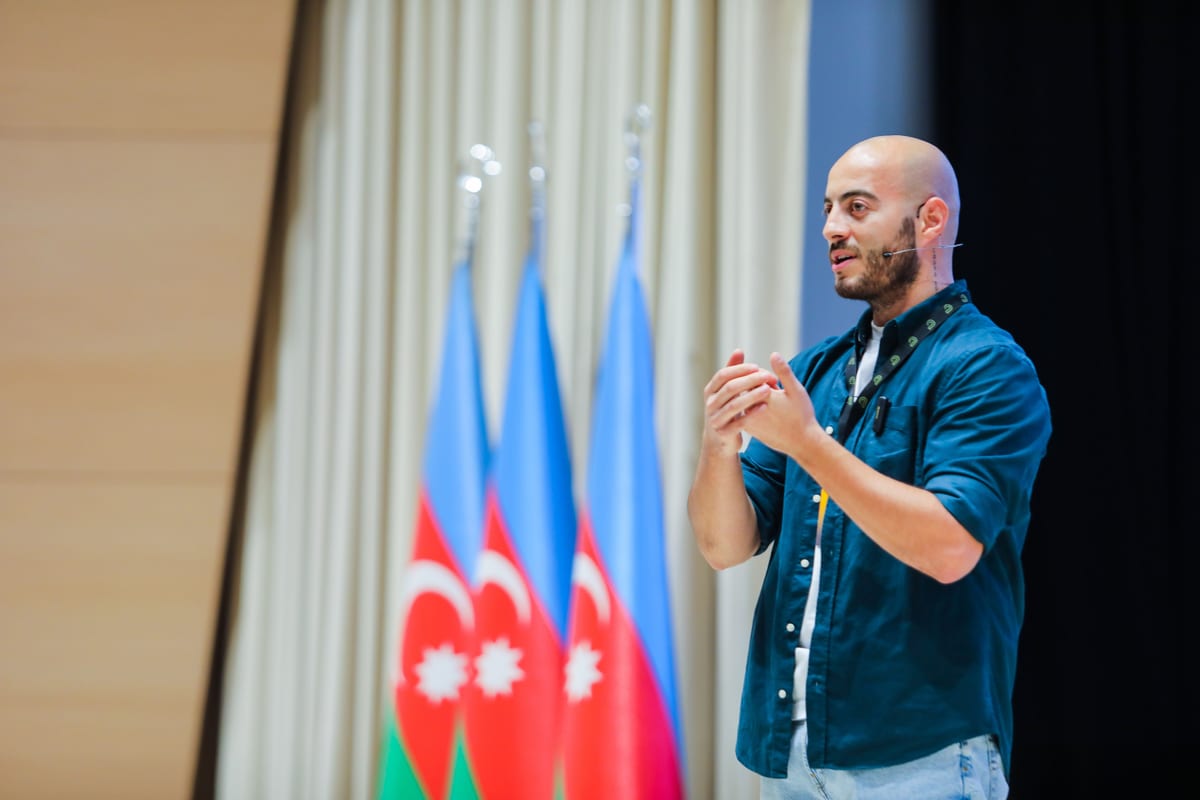 Man speaking at an event in Baku, Azerbaijan, with Azerbaijani flags in the background.