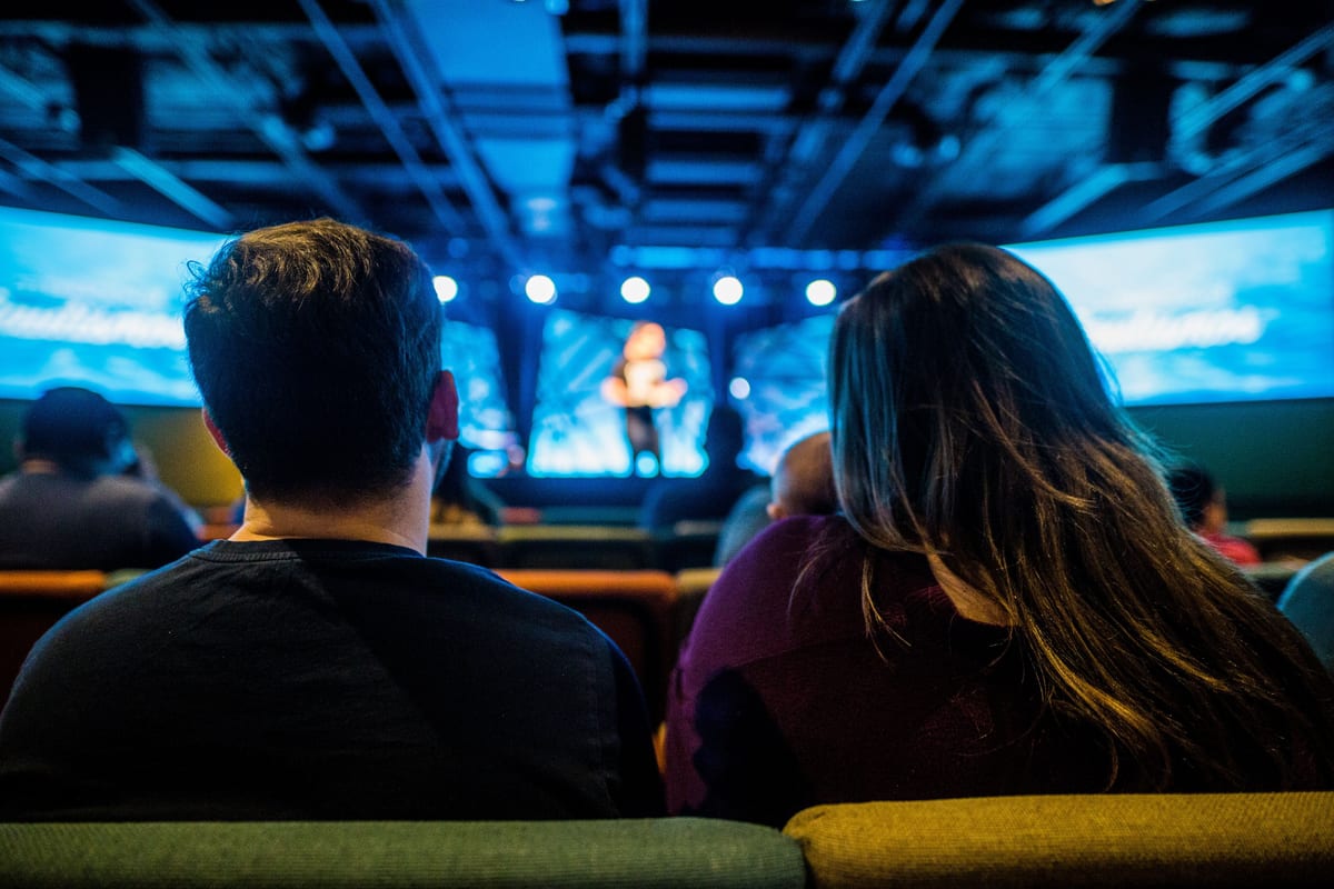 Two adults attending a conference, viewed from behind, with a speaker on stage.