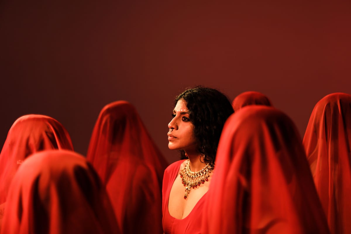 Dramatic portrait of a woman in a red blouse with veiled figures around her.