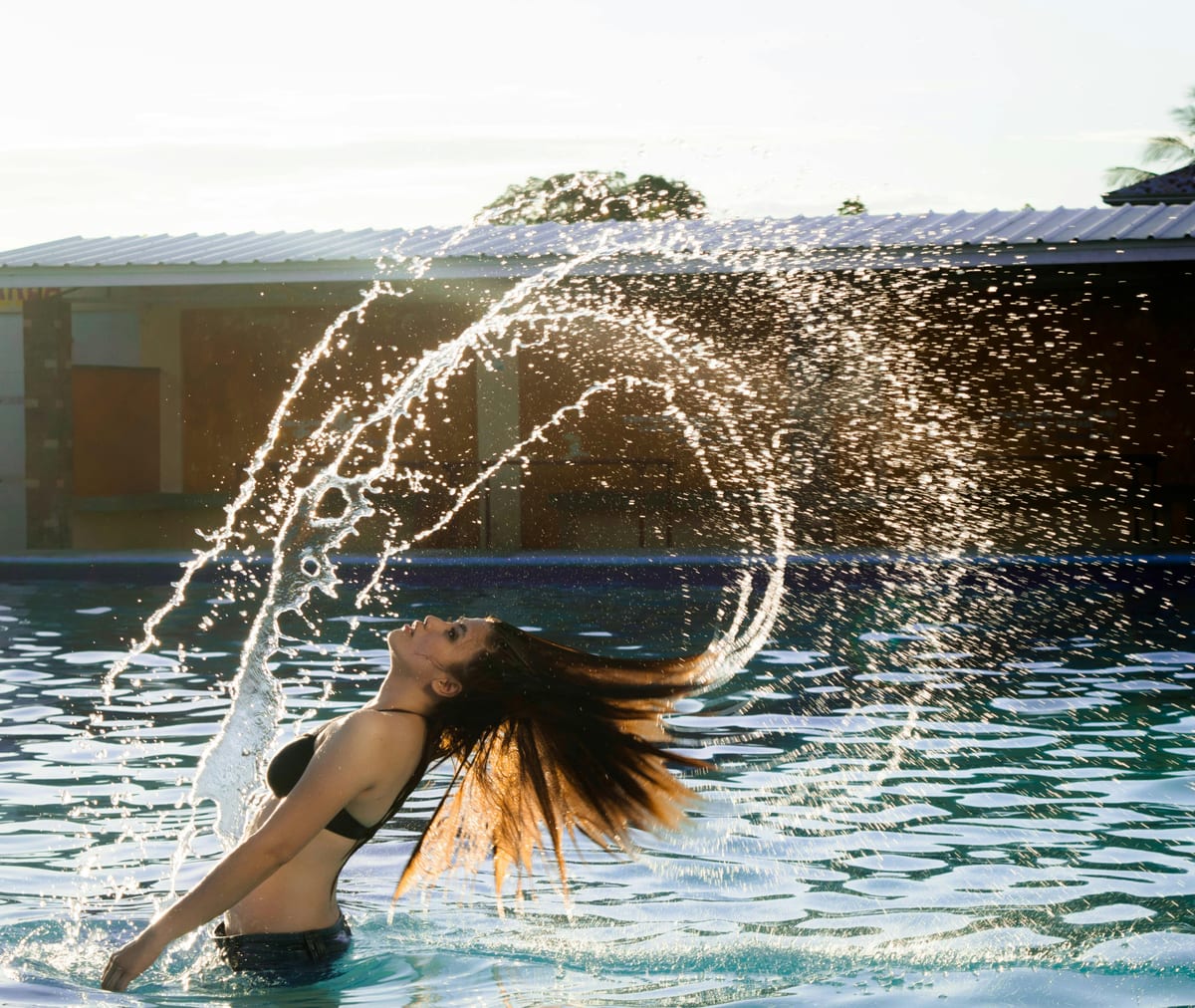 A young woman flips her hair in a swimming pool, creating a splash of water.