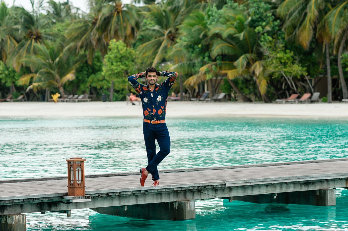 Man in floral outfit poses on a pier in tropical Maldives setting.