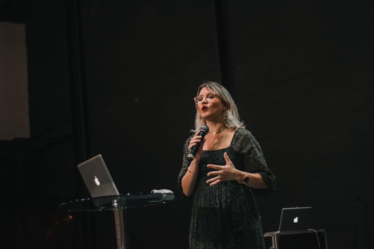 A confident woman delivers a speech on stage with a microphone and laptop.