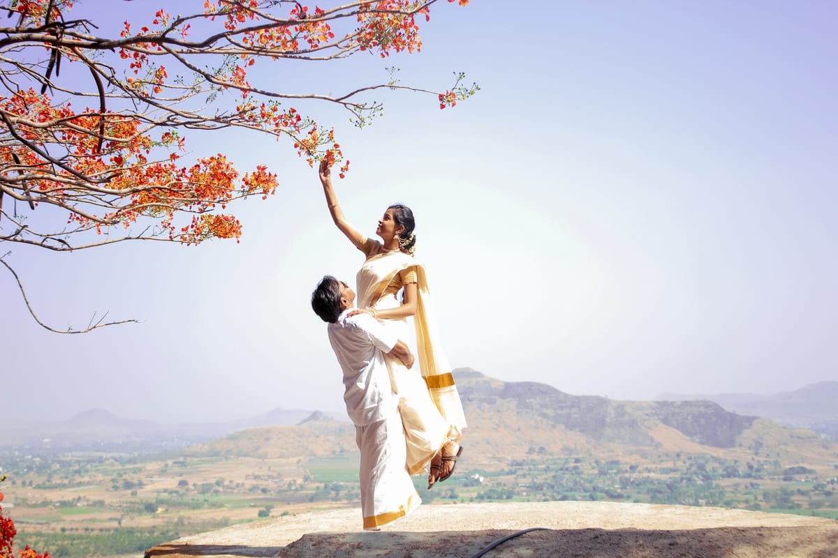 A joyful Indian couple in traditional attire celebrates love under blooming branches in a sunny landscape.
