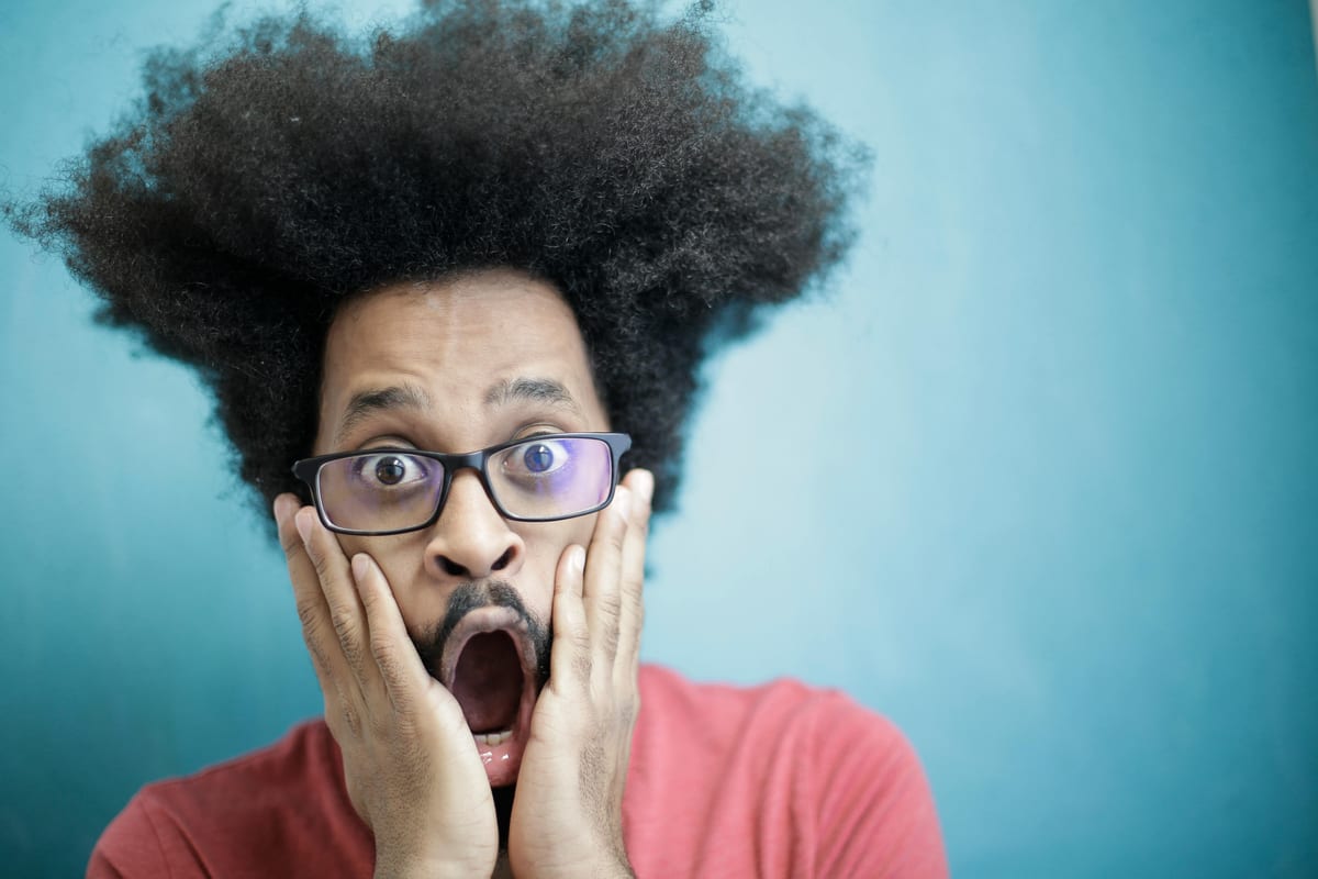 Close-up portrait of a surprised man with afro hairstyle and glasses against a blue background.