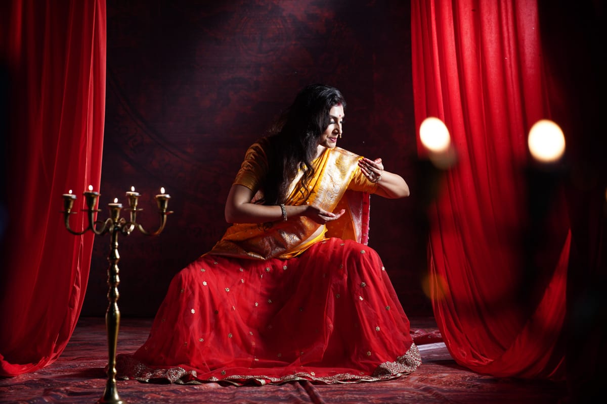 A woman performs a traditional dance in a vibrant red saree, surrounded by red drapes and candles.