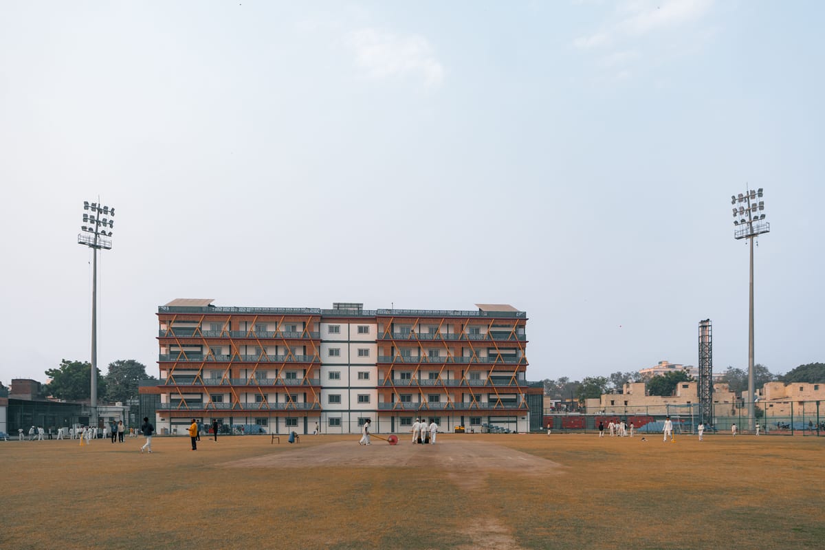 Cricket match in progress at a sports field in Delhi, India, with players and spectators under clear skies.