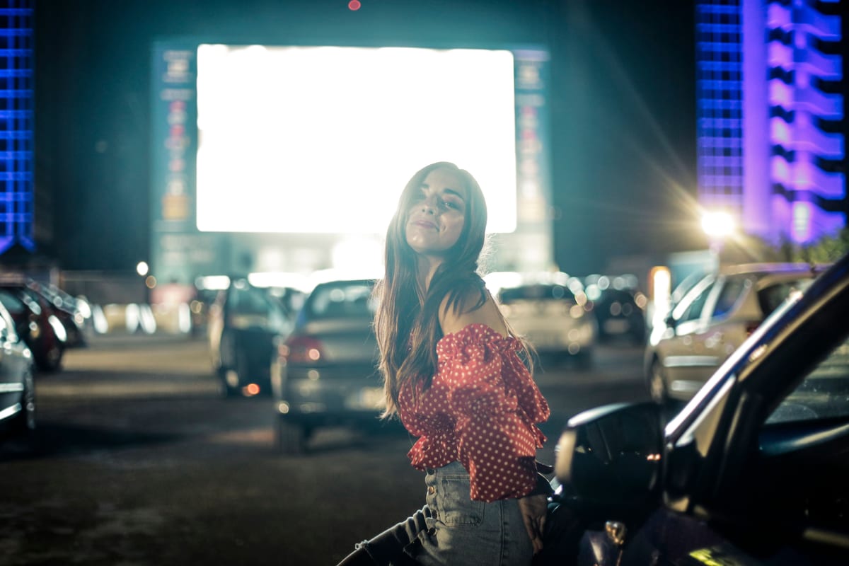 Beautiful female with dark hair looking at camera while standing near automobile on street with cars and illuminated screen on blurred background at twilight