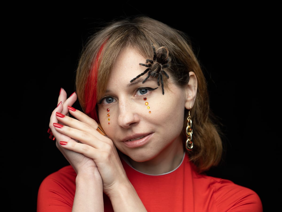 Calm young woman in red clothes with artistic makeup and big spider on face looking at camera pensively against black background