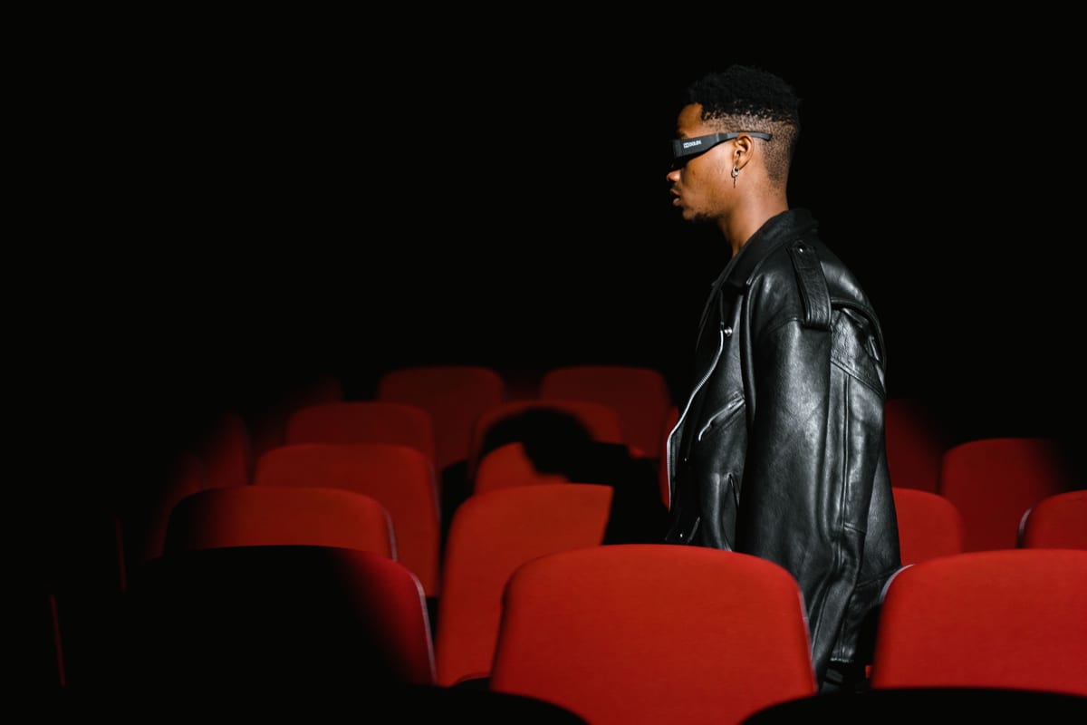 A man wearing glasses walks through a dimly lit movie theater with red seats.