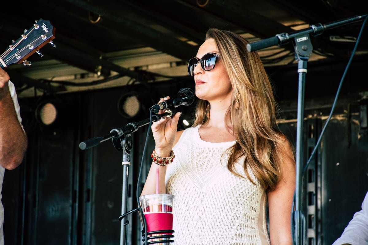 A woman in sunglasses sings on stage during an outdoor concert, exuding confidence and style.