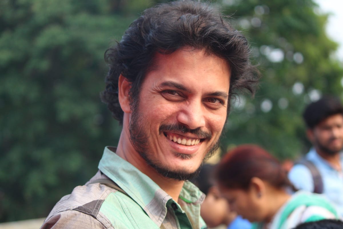 Cheerful man smiling outdoors in West Bengal, India with blurred people in the background.