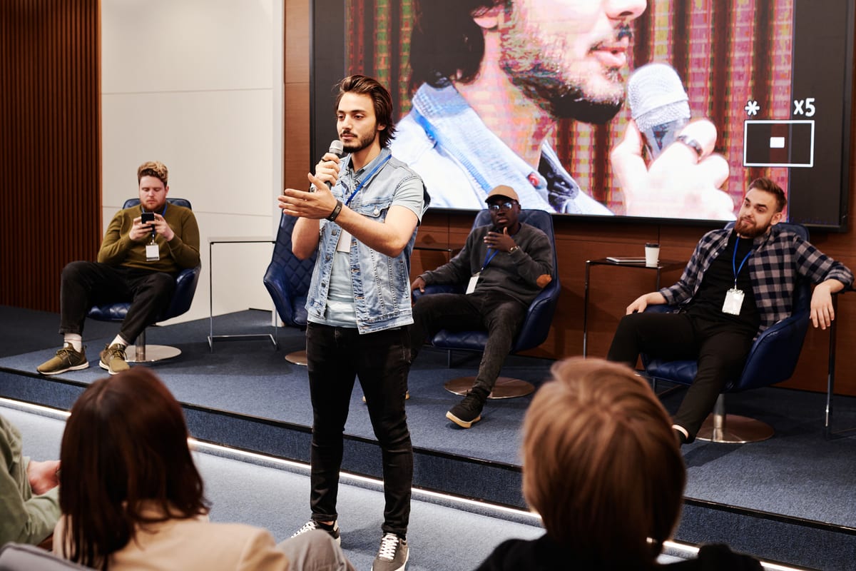 Speaker holds microphone, engaging audience at an indoor business conference with a panel in the background.