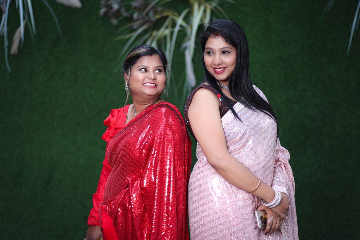Two Indian women in traditional sarees smiling and posing against a green background.