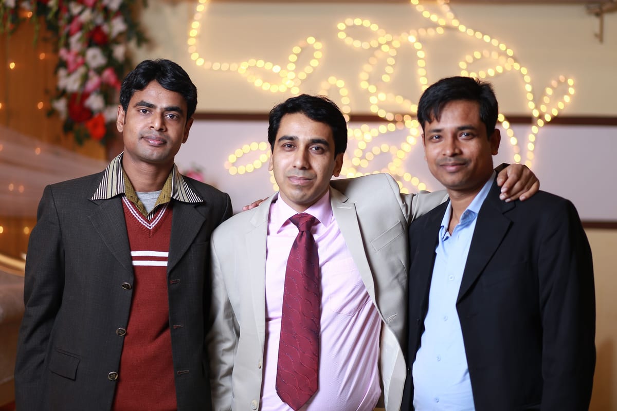 Three men in formal attire smiling together at an indoor event with festive lighting.