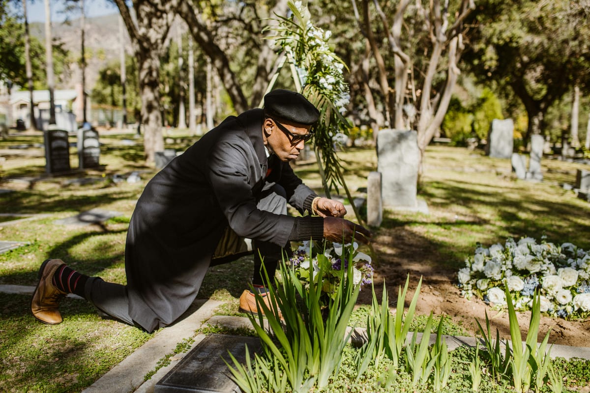 A man kneeling and attending to a grave at a sunlit cemetery.