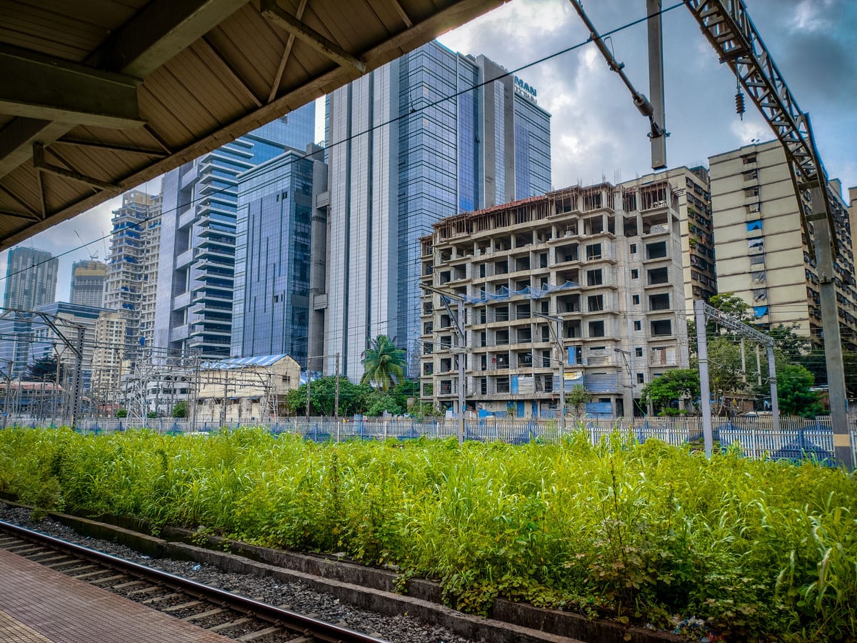 Modern skyscrapers in Mumbai viewed from a railway platform with lush greenery in the foreground.